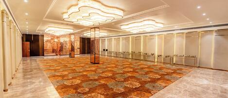 Un'ampia hall d'albergo con un grande tappeto decorato, una colonna centrale ornamentale e una fila di armadietti a destra.