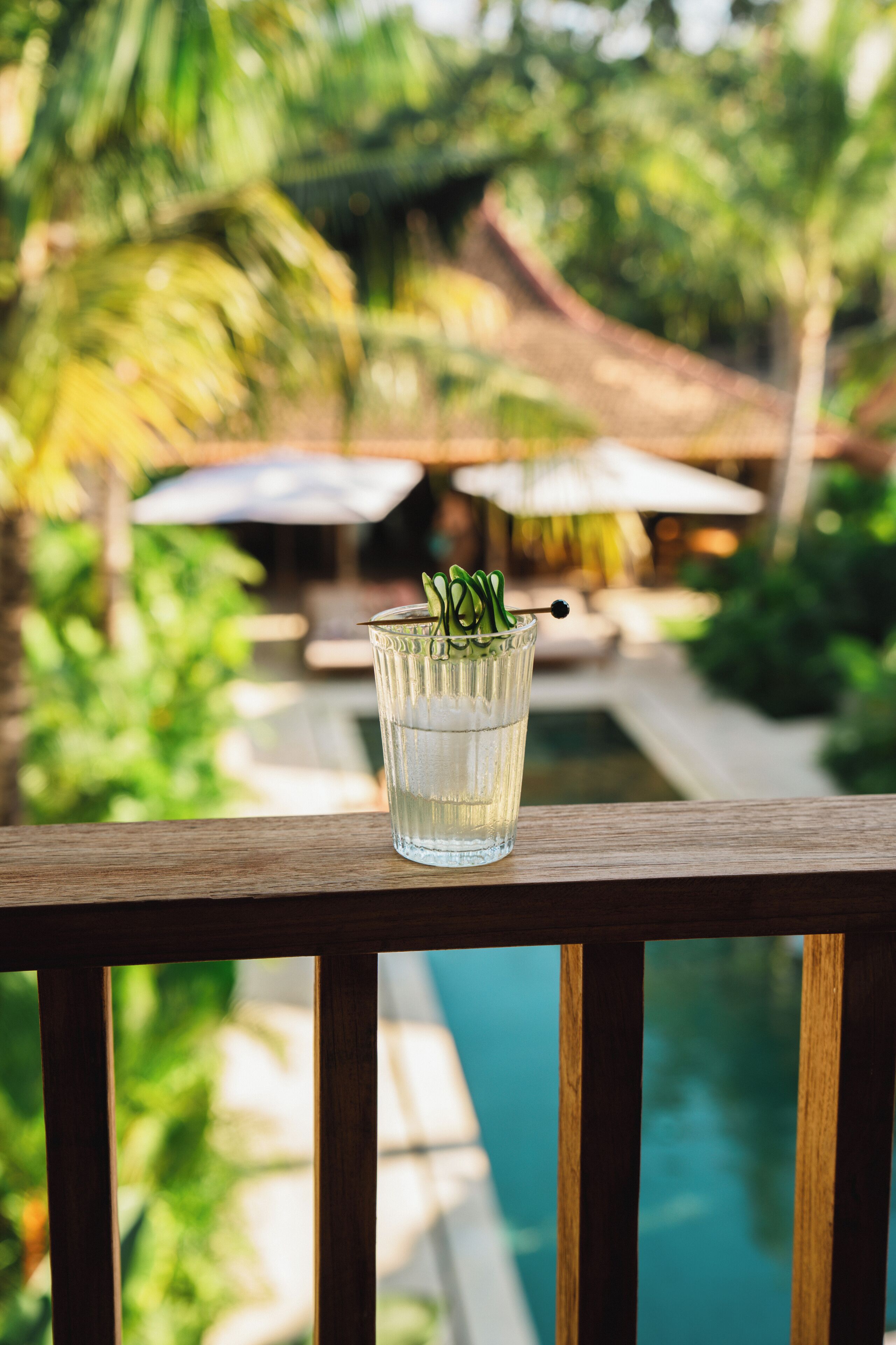 Un vaso de agua con una planta sobre una terraza de madera con vistas a una piscina y abundante vegetación.