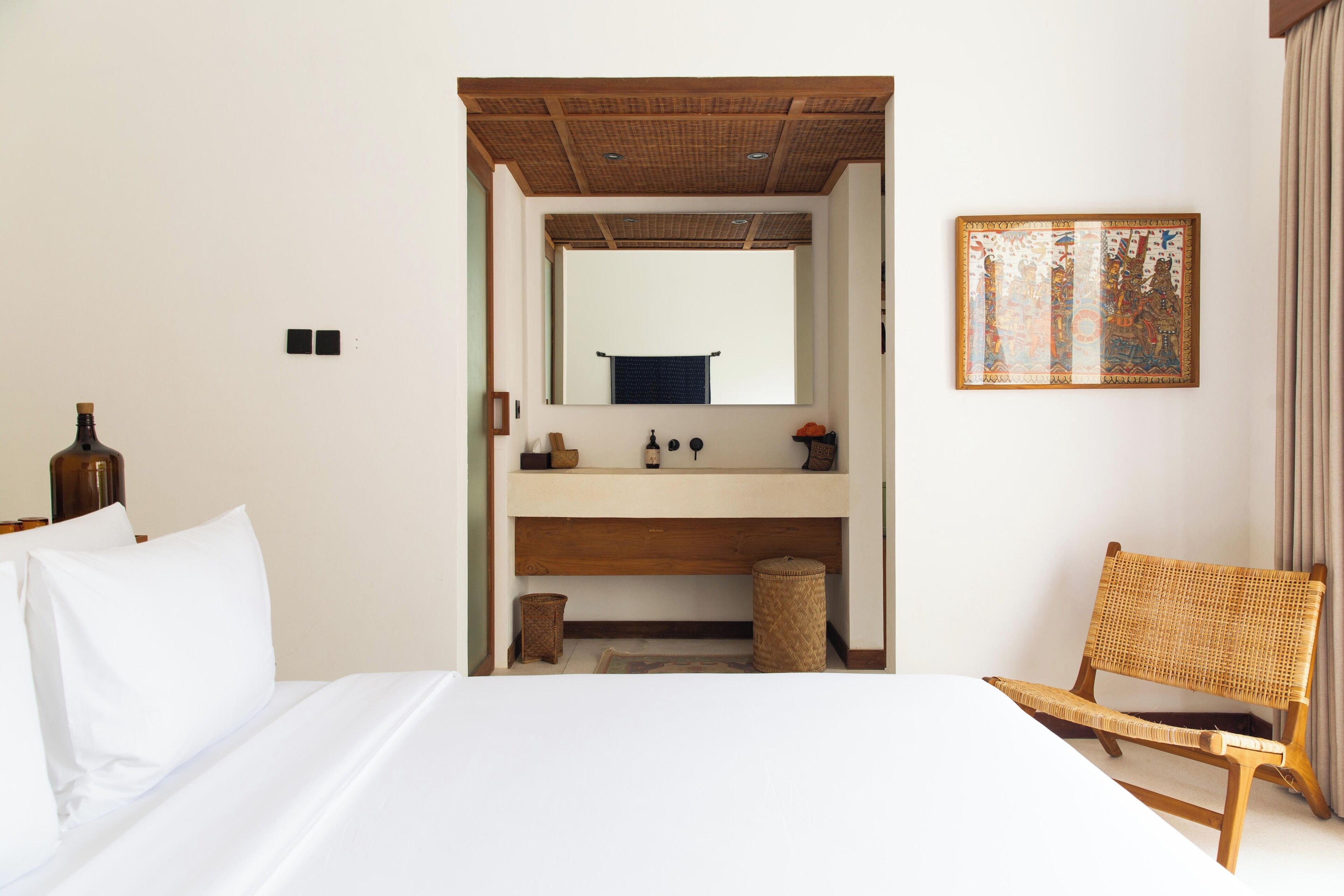 Habitació d'hotel amb llit, cadira de fusta, bany amb rentapla i mirall, i una obra d'art emmarcada a la paret.