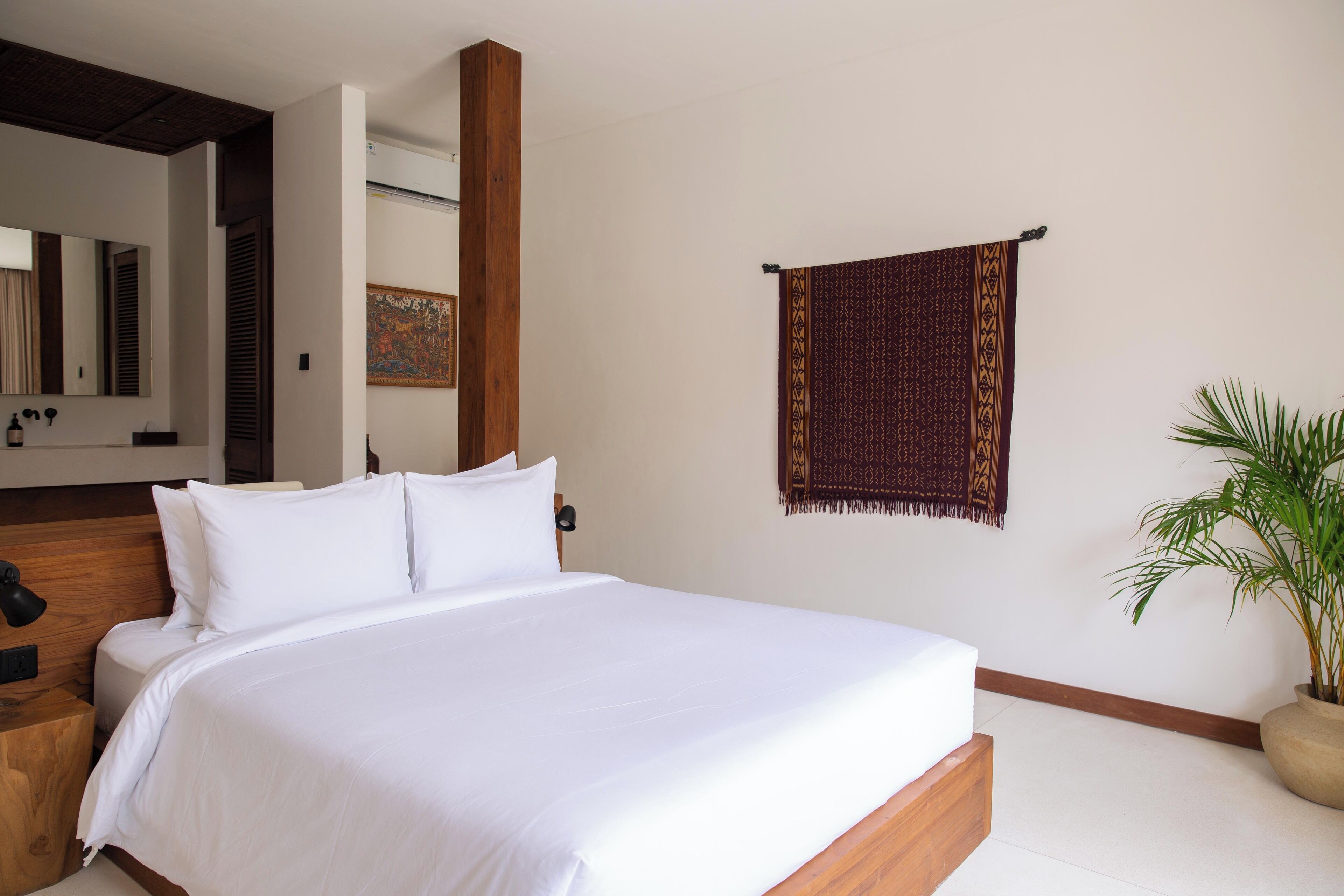 A neatly made bed with white linens, a wooden headboard, a wall-mounted tapestry, and a potted plant.