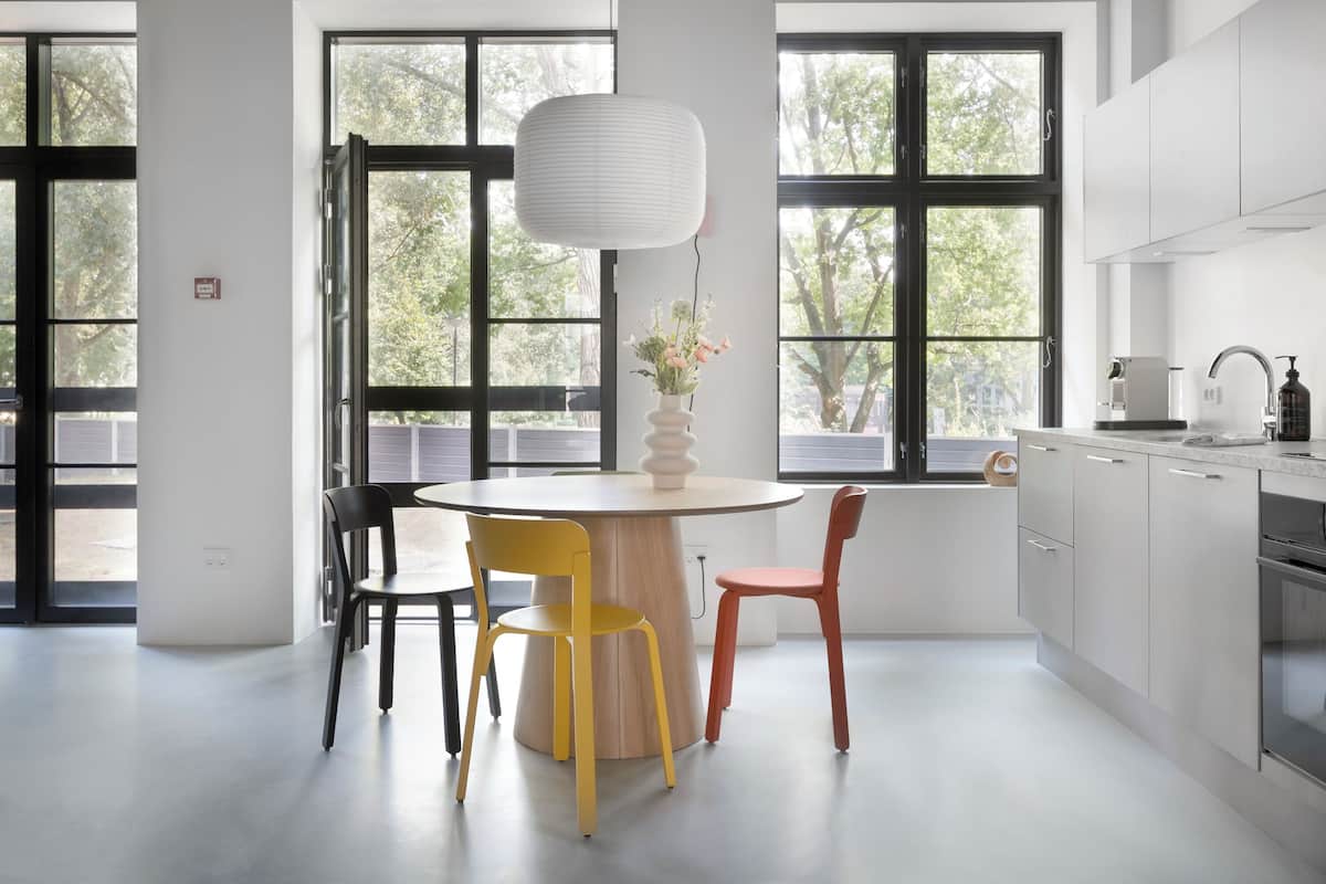 Een moderne keuken met een ronde eettafel, twee stoelen, een vaas met bloemen en grote ramen met uitzicht op bomen.