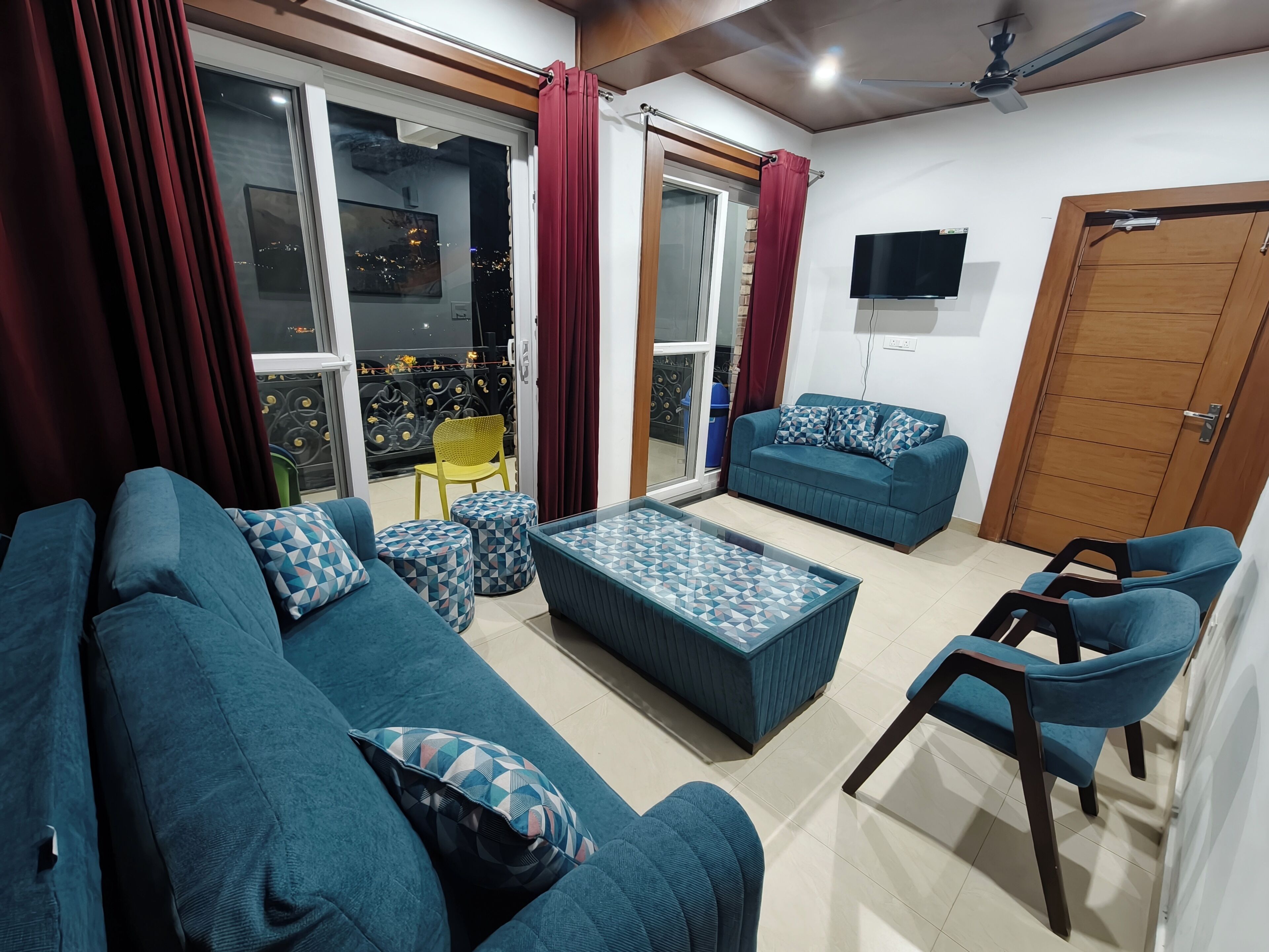 A living room with a sofa, ottoman, and chairs, a TV, and a sliding glass door to a balcony.