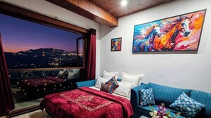 Habitación de hotel con una cama grande, un sofá azul, una mesita de centro de cristal, un cuadro y vistas a un paisaje urbano nocturno.