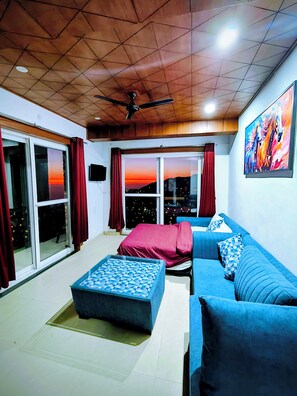 A room with a wooden ceiling, a blue sofa, a red bed, and a view of the sunset through the windows. - Opal Villas (Mussoorie)