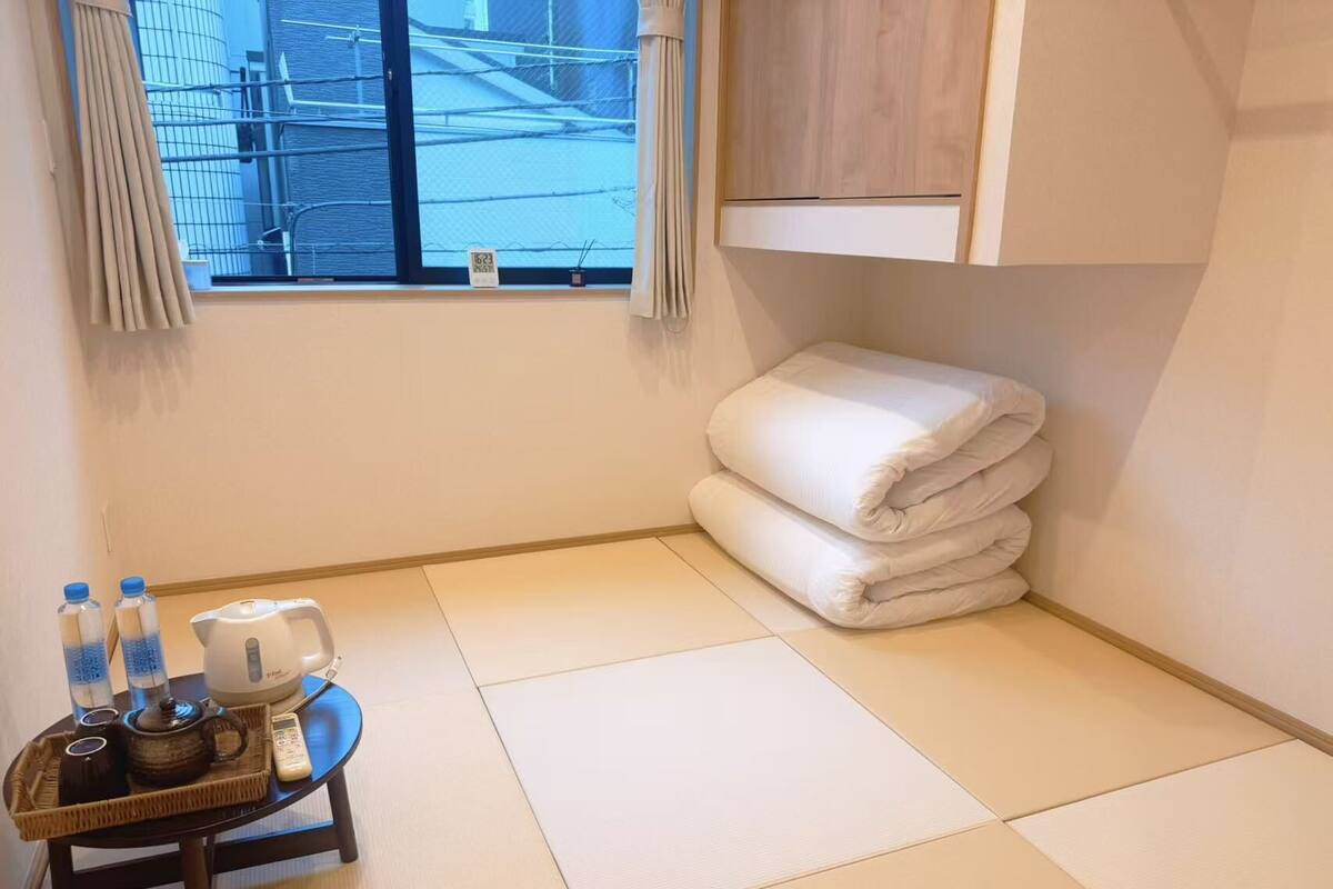 A hotel room with tatami flooring, a small table with tea set, a stack of towels, and a window with a view of buildings.