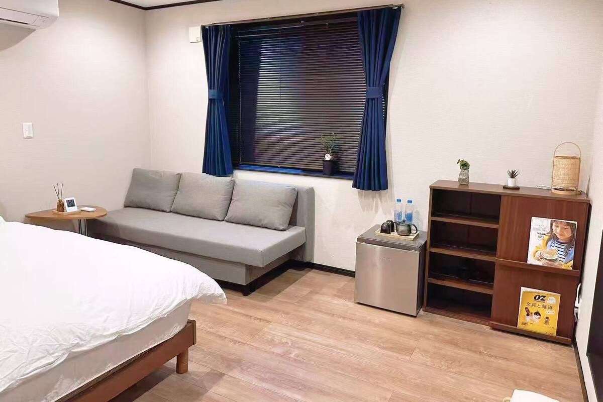 A hotel room with a bed, a sofa, a small table with a plant, a shelf with books and a magazine, and a window with curtains.