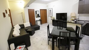 Una sala moderna con un sofá negro, una mesa de comedor con sillas, un televisor de pantalla plana y una cocina con armarios y encimera.