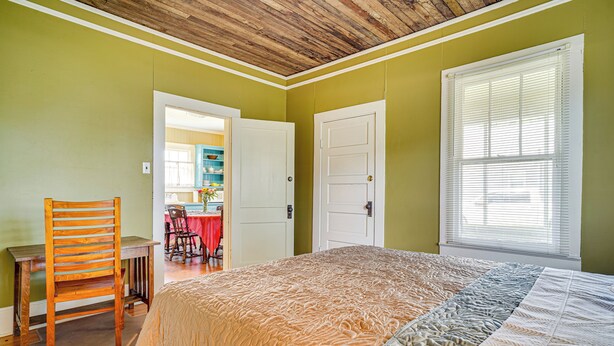 A bedroom with a wooden ceiling, a bed with a patterned bedspread, a wooden chair, and a door leading to a dining area. - Walkable Dtwn Lafayette Home w/ Screened Porch (Lafayette)