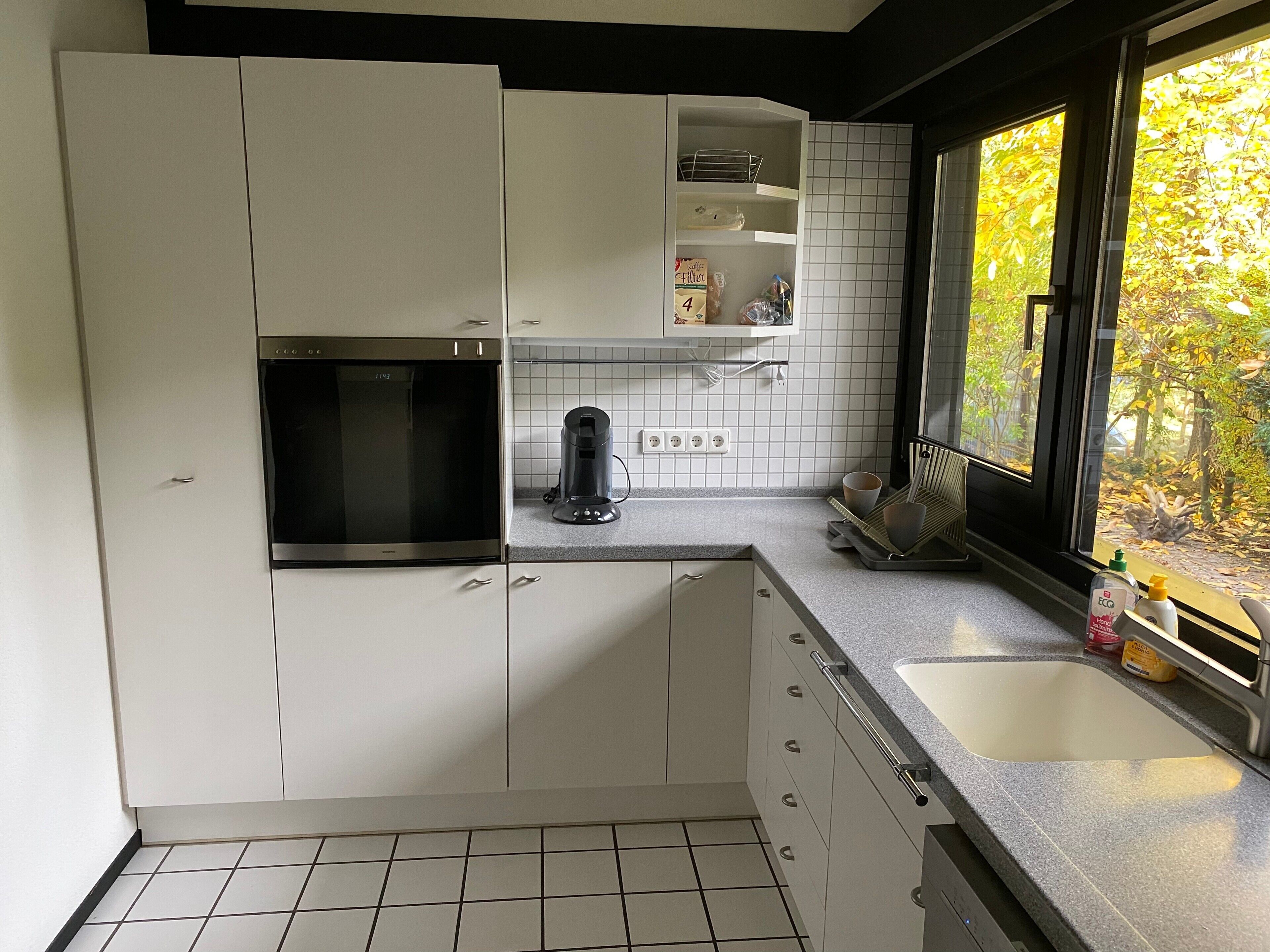 A modern kitchen with white cabinets, a built-in microwave, a coffee maker, and a window with a view of trees.