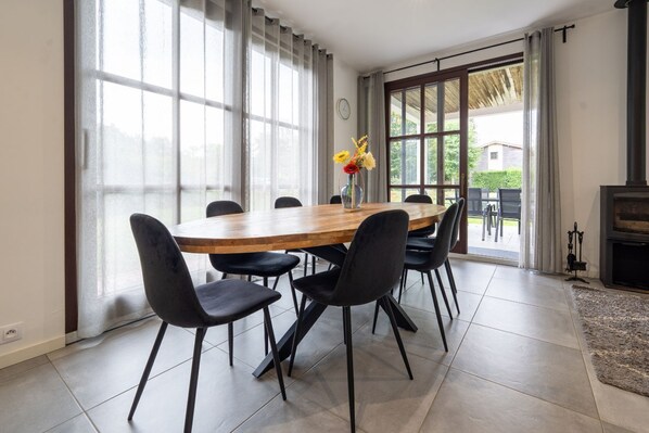 A dining area with a wooden table and black chairs, large windows with curtains, and a view of the outdoors.