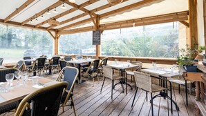 Un'area pranzo all'aperto con tavoli e sedie in legno, ampie finestre e una struttura a soffitto in legno.