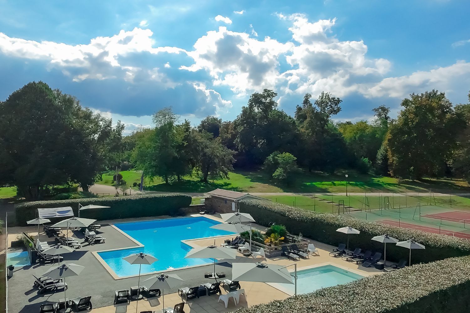 A pool area with multiple sun loungers and umbrellas, surrounded by well-maintained hedges and trees.