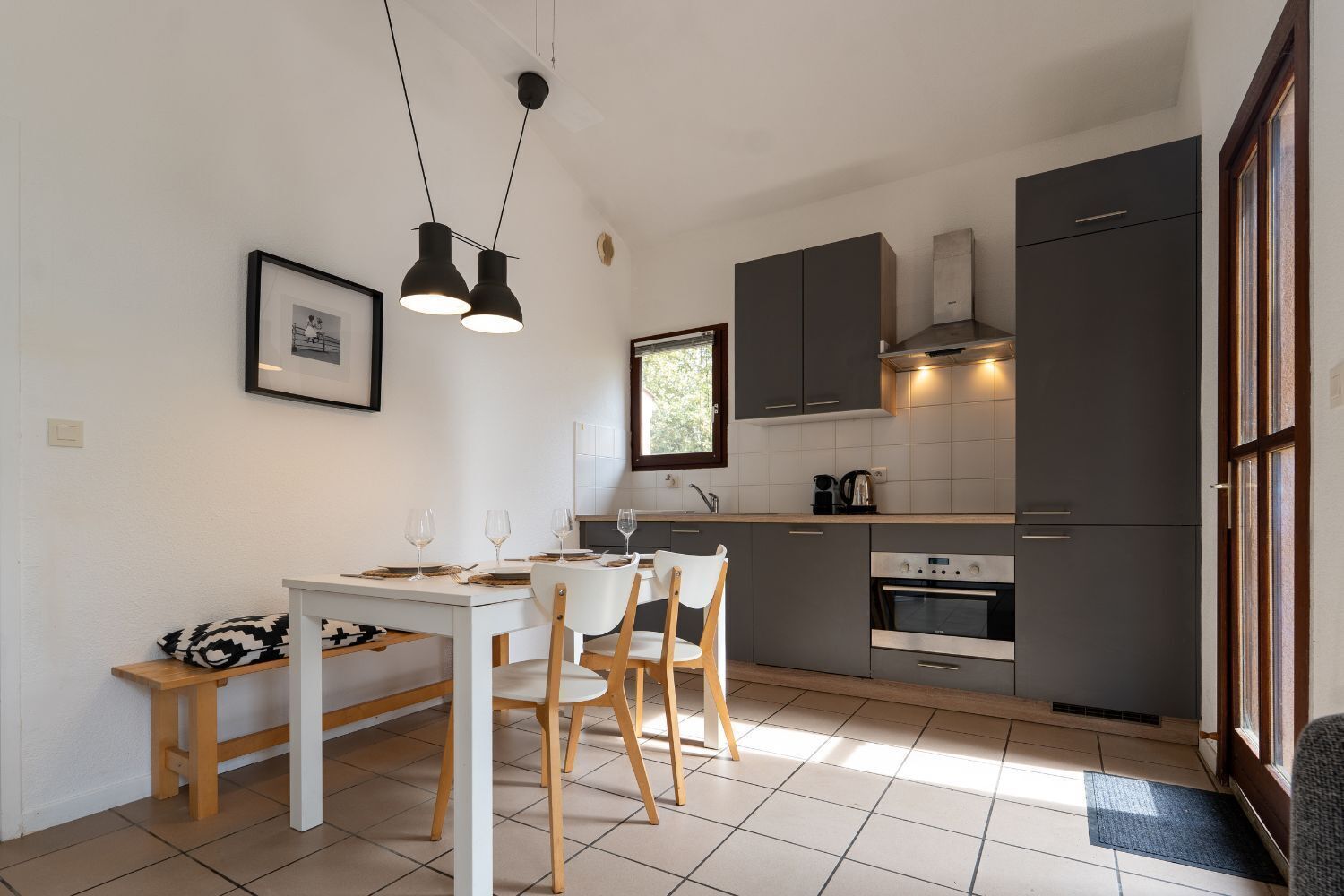 A compact kitchen with a dining area, featuring a white table, two chairs, a small wooden side table, and a framed picture on the wall.