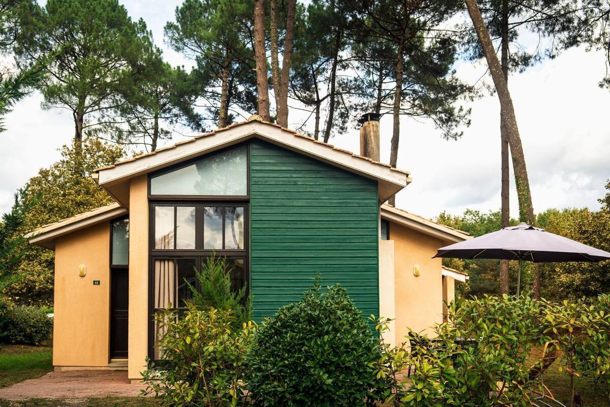 A small, single-story house with a green exterior and a beige roof, surrounded by lush greenery and trees.