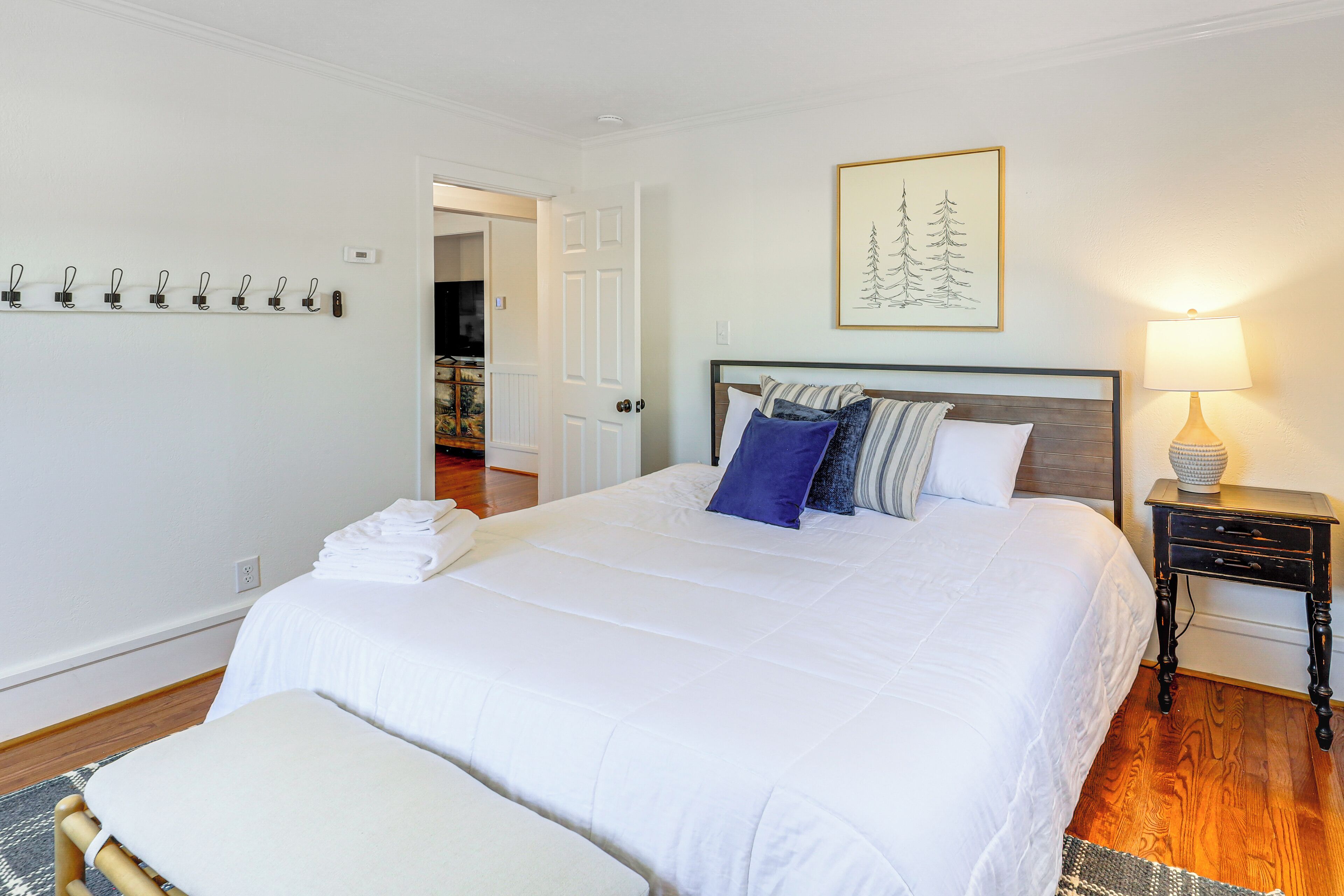 A neatly made bed with a white comforter and several pillows, a bedside table with a lamp, and a wall-mounted coat rack with several hangers.