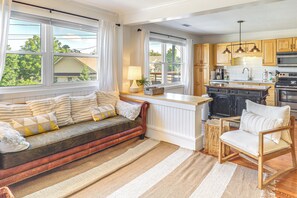 Ein gemütliches Wohnzimmer mit Sofa und Sessel, ein Küchenbereich mit Holdekor-Schränken und Geräten sowie große Fenster mit Vorhängen.