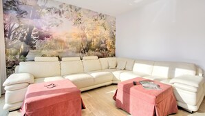Ein modernes Wohnzimmer mit einem Ecksofa, zwei Poufs und einem großen Wandbild mit Waldmotiv.