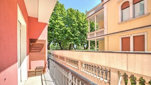 Ein Balkon mit Metallgeländer, einer Holzbank und Blick auf Bäume sowie ein Gebäude mit Balkon.