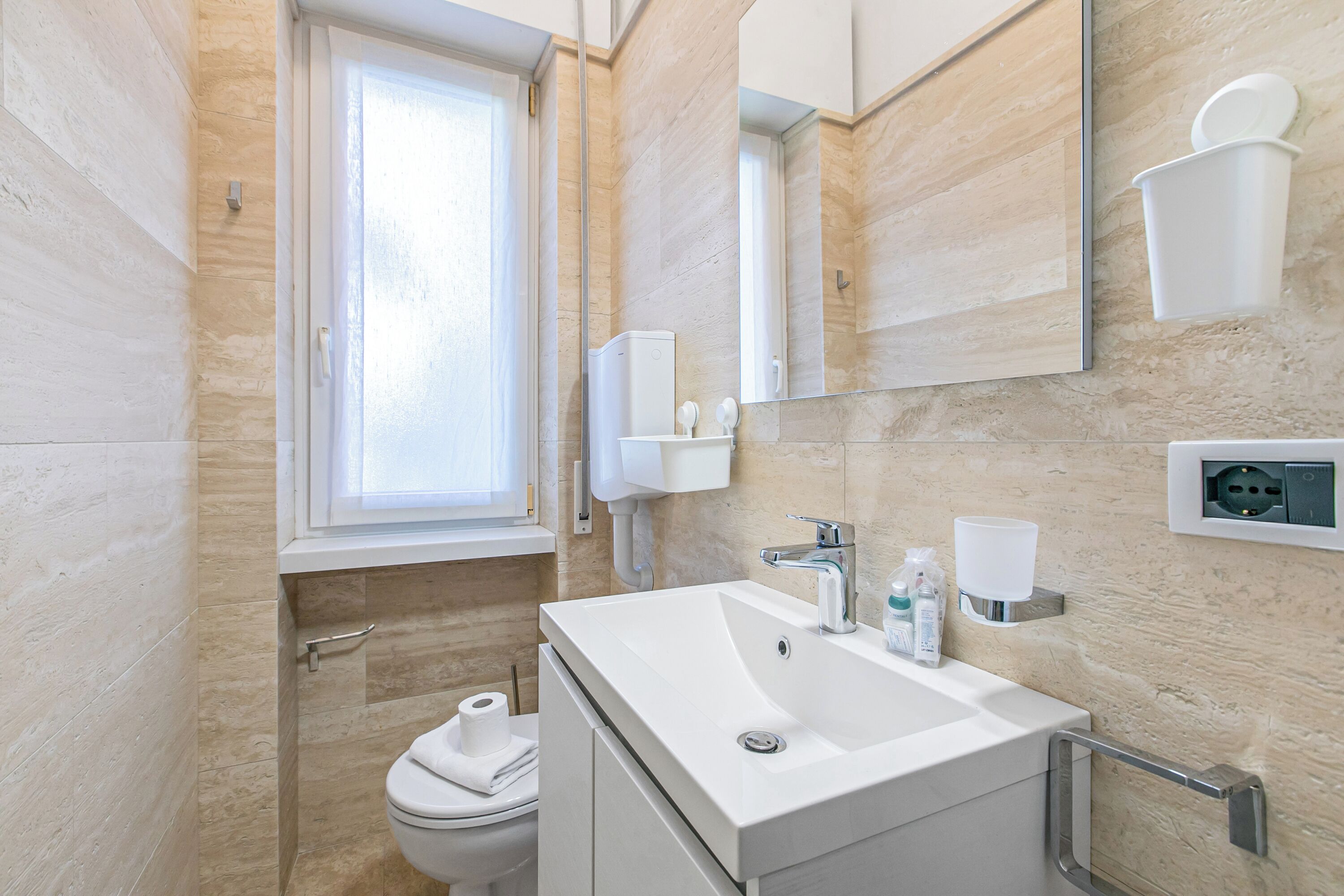 A bathroom with a toilet, sink, mirror, window, and towel dispenser.