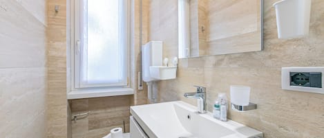 A bathroom with a toilet, sink, mirror, window, and towel dispenser.