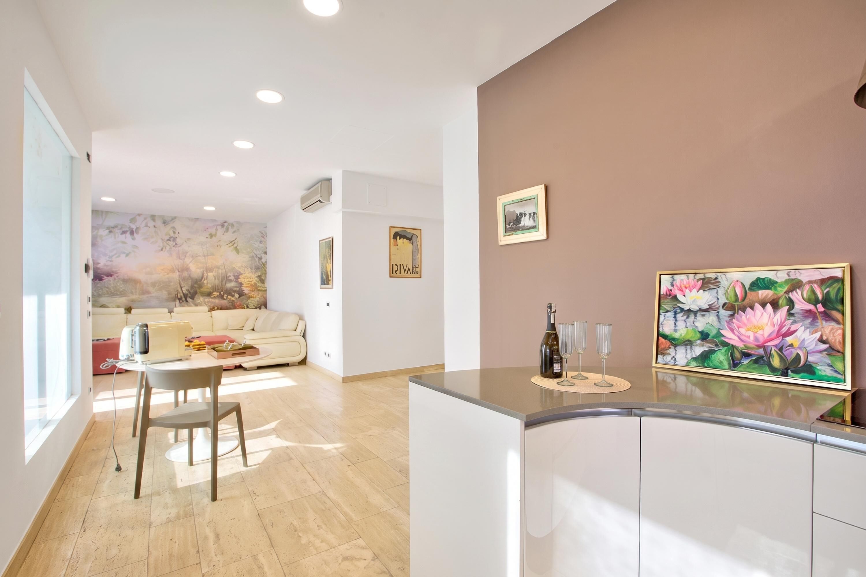 A modern kitchen with a dining area, featuring a wooden floor, white cabinetry, and a large painting of flowers on the wall.