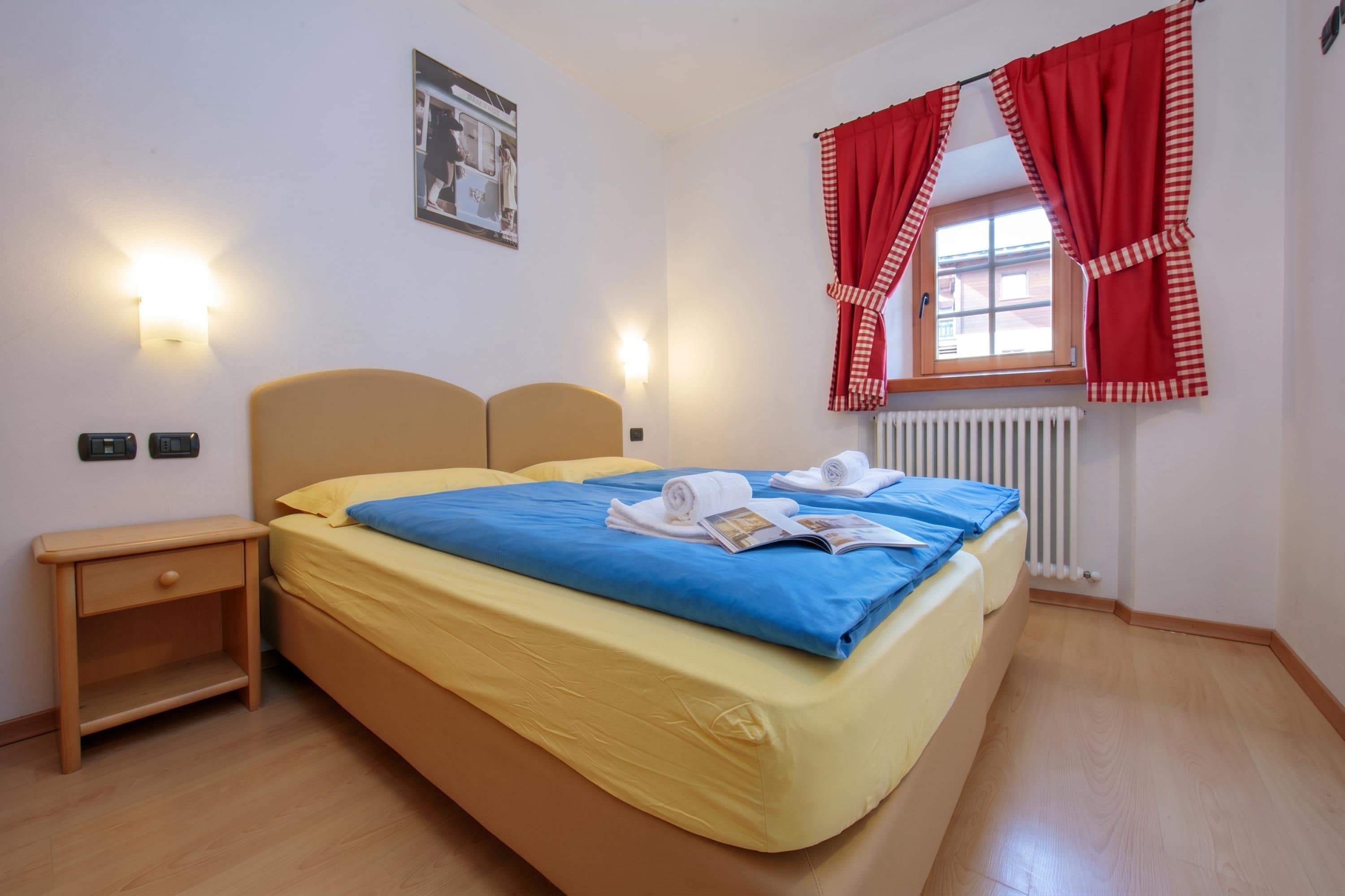 Une chambre d’hôtel avec un lit, des tables de chevet, une fenêtre avec des rideaux rouges et un lampadaire fixé au mur.