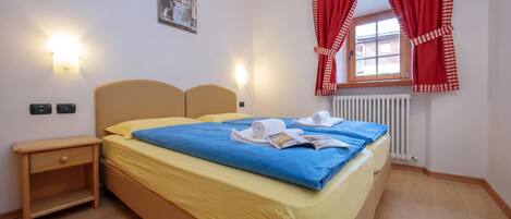A hotel room with a bed, bedside tables, a window with red curtains, and a wall-mounted light.