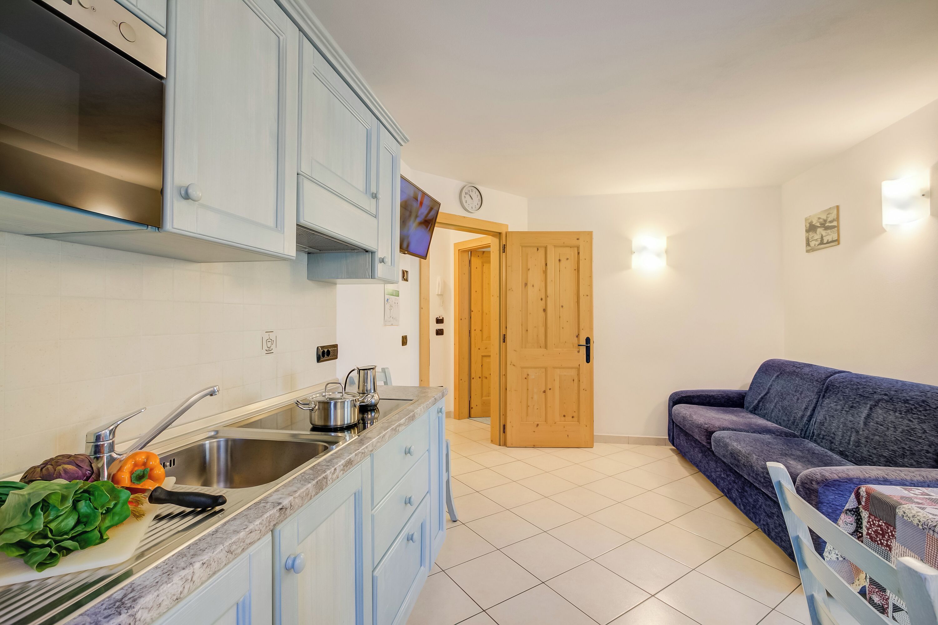 Een keuken met een spoelbak, kastjes en een aanrecht met groenten. In de woonkamer staat een houten deur en een blauwe bank.