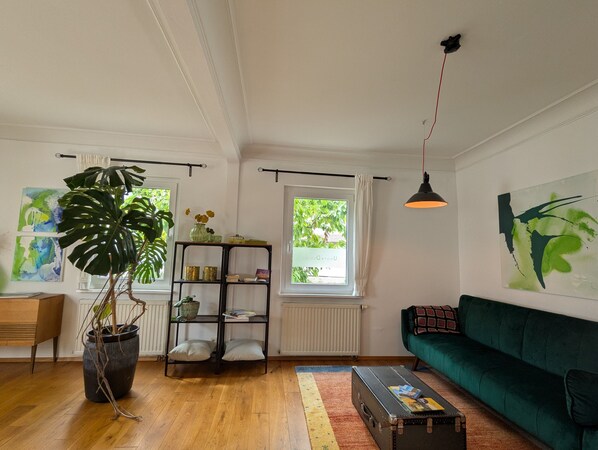 A living room with a green sofa, a wooden floor, a black lamp, a bookshelf, and a large plant.