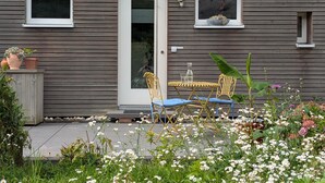 Una casa de fachada gris, con una puerta blanca y un ventanal pequeño.  Una mesa y dos sillas se encuentran en el exterior, rodeadas por un jardín con diversas plantas y flores.