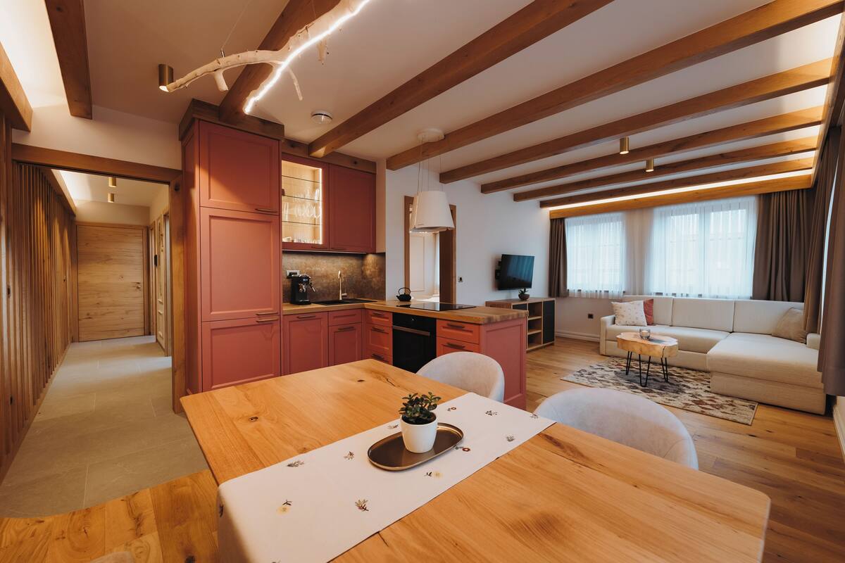 A modern kitchen with wooden beams, a dining area with a table and chairs, and a living room with a sofa and a TV.