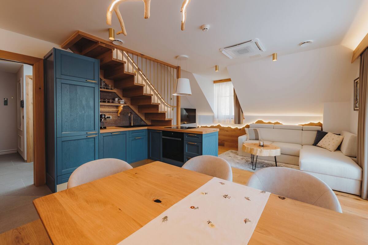 A modern kitchen with blue cabinets, a wooden dining table, and a staircase leading to a loft bedroom.