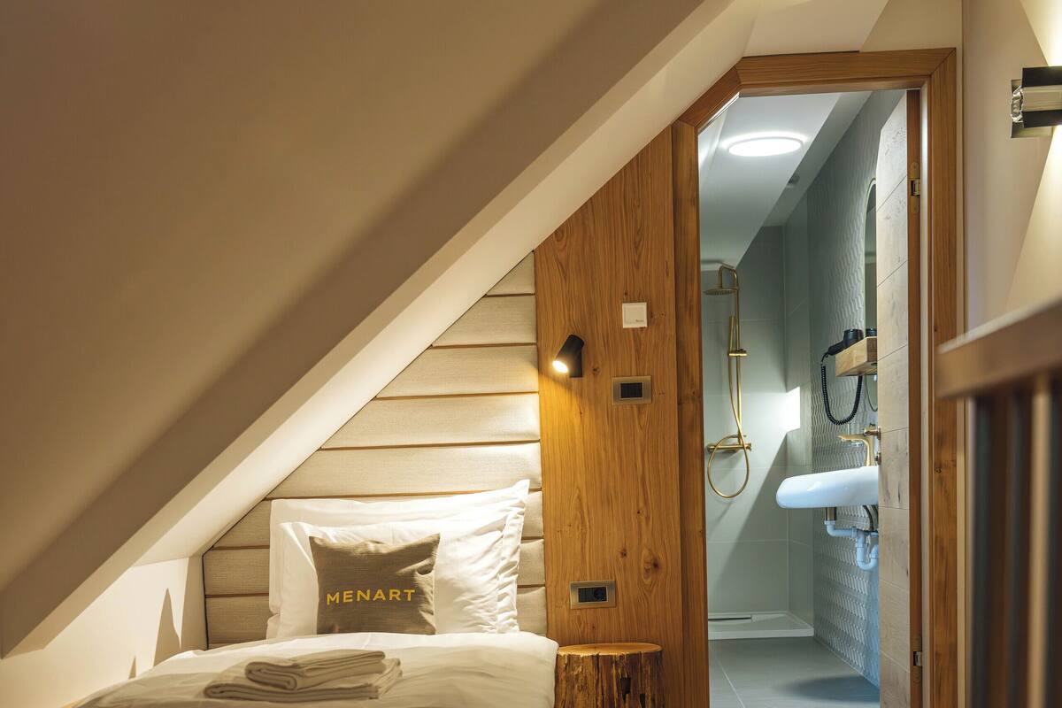A bedroom with a sloped ceiling, wooden flooring, a bed with a pillow labeled "MENART," a bedside table, and a view into a bathroom with a sink and shower.