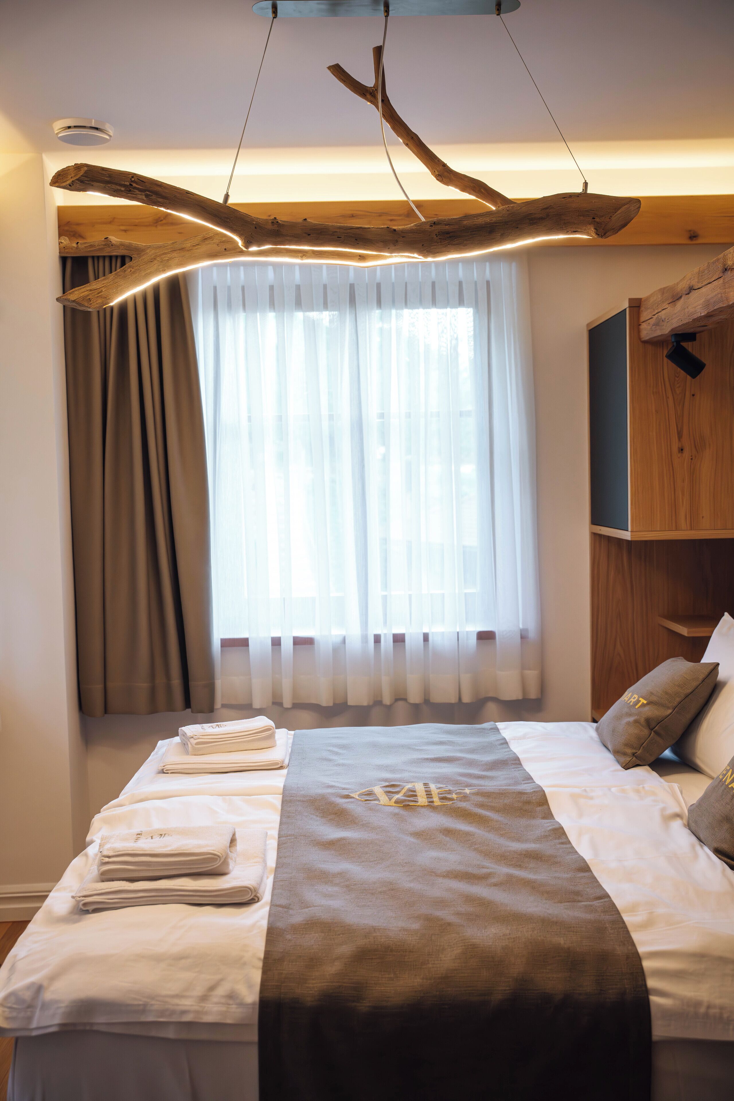 Habitación de hotel con cama, techo de madera decorativo, ventana con cortinas y repisa con almohadas.
