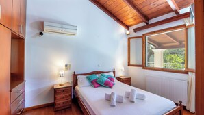 Un dormitorio con techo de madera, una cama con ropa blanca y almohadas coloridas, una mesita de noche con lámpara, y una ventana con vista a follaje.