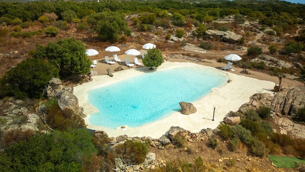 A pool surrounded by white lounge chairs and umbrellas, set in a natural landscape with rocky terrain and sparse vegetation.