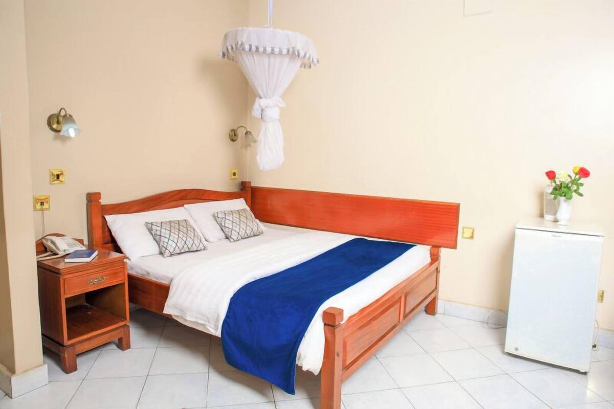 A bedroom with a wooden bed, a nightstand, a bedside lamp, a small refrigerator, and a hanging mosquito net.