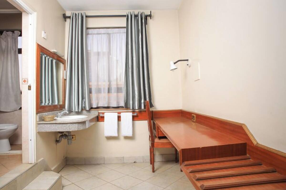 A small bathroom with a sink, mirror, and window with curtains. There is a wooden bench and a towel rack.