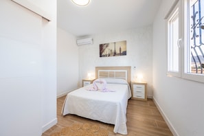 A bedroom with a bed, two bedside tables, a wall-mounted air conditioner, a cityscape painting, and a window with a view. - Apartment 'La Palmera' with Private Terrace and Air Conditioning (El Nazareno)