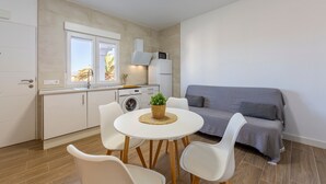 Une cuisine compacte avec des meubles blancs, un four à micro-ondes et un lave-linge. On y trouve une table ronde avec des chaises et un canapé gris dans l’espace salon.