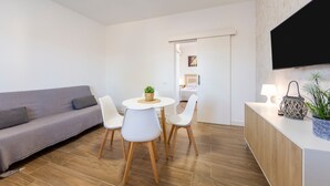 Un salon moderne avec un canapé gris, une table à manger ronde entourée de chaises, un téléviseur fixé au mur et un meuble en bois agrémenté d’une plante.