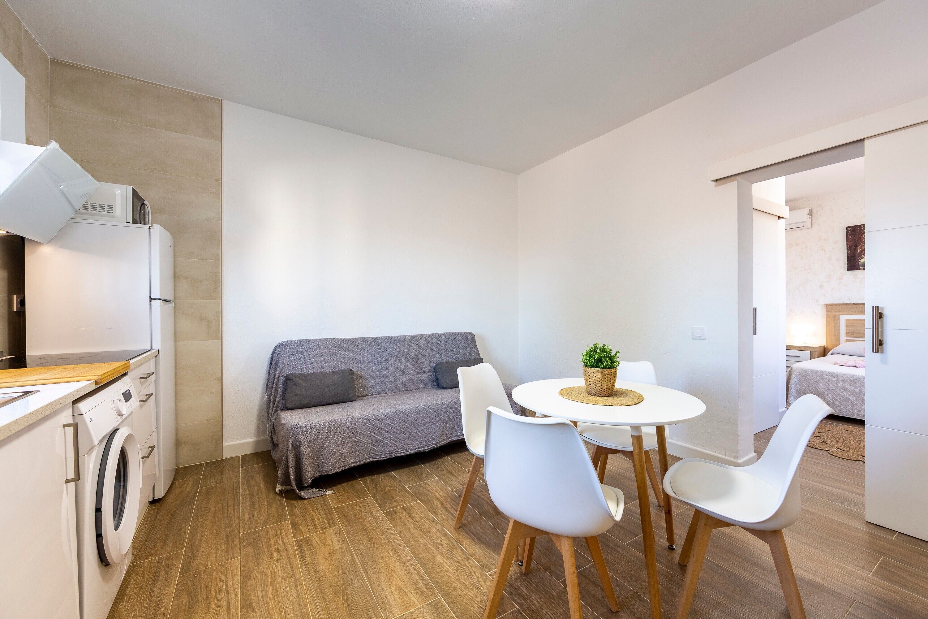 Een knus woongedeelte met een wasmachine, een grijze bank, een eettafel met stoelen en een kleine keuken.