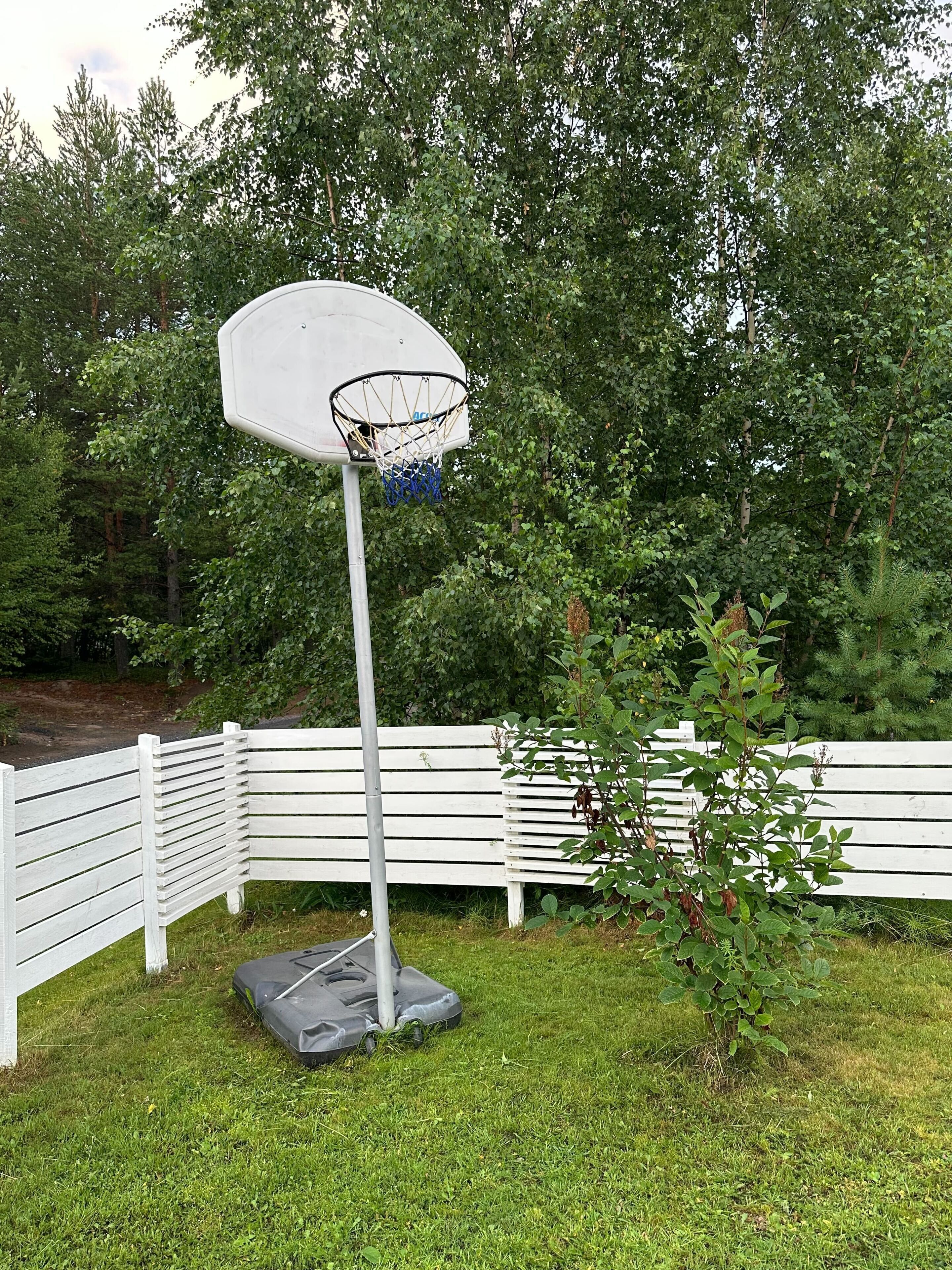 A basketball hoop in a grassy area with a white fence and trees in the background.