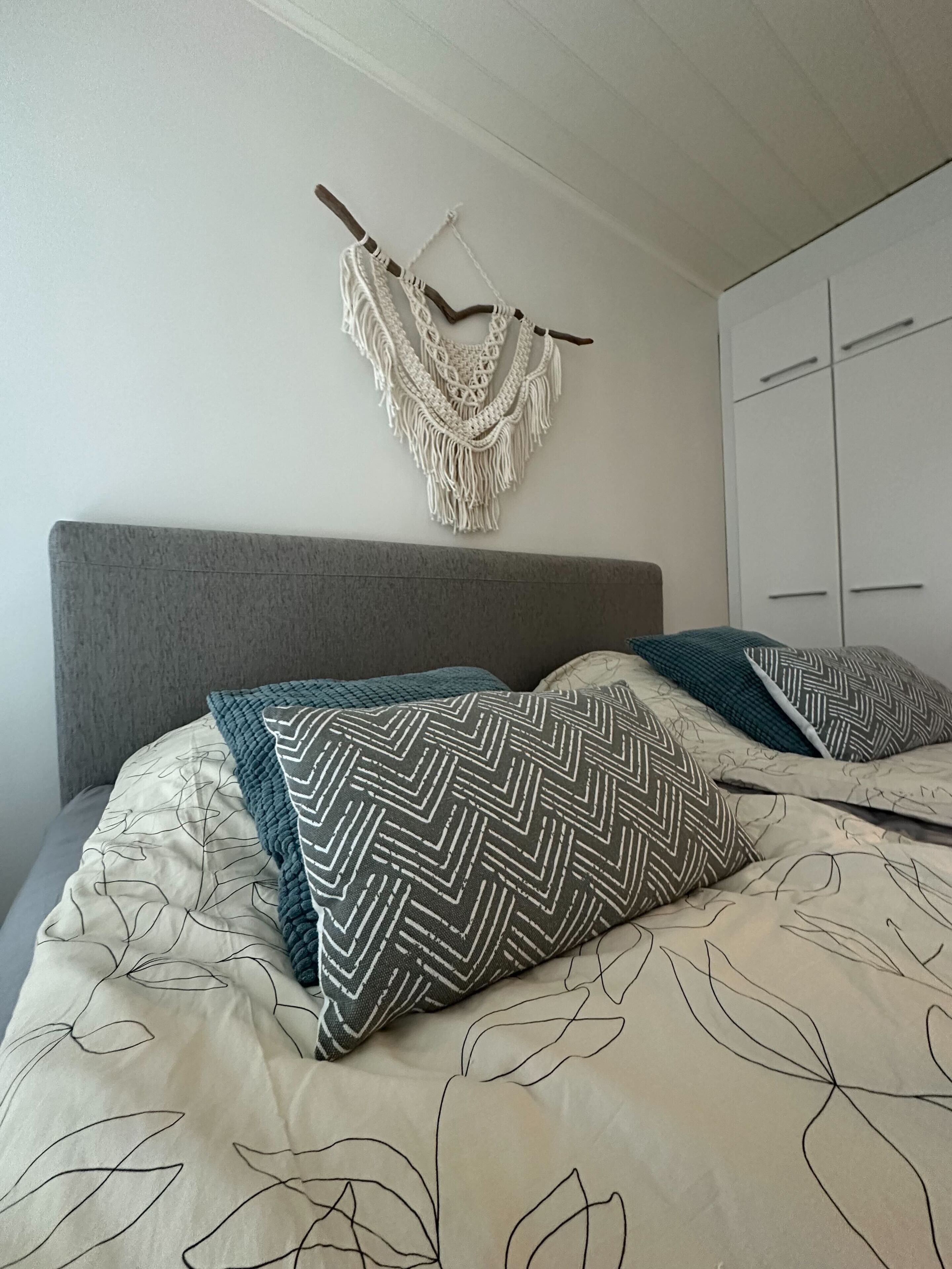 Un dormitorio con cama, cabecera gris, armario blanco y un colgador de macramé en la pared.