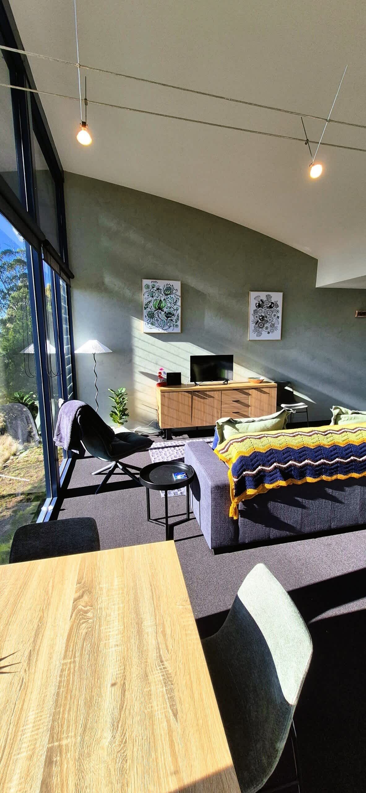 Een moderne woonkamer met een bank, een eettafel en een televisie. Twee kunstwerken hangen aan de muur, en een raam biedt uitzicht op het landschap.