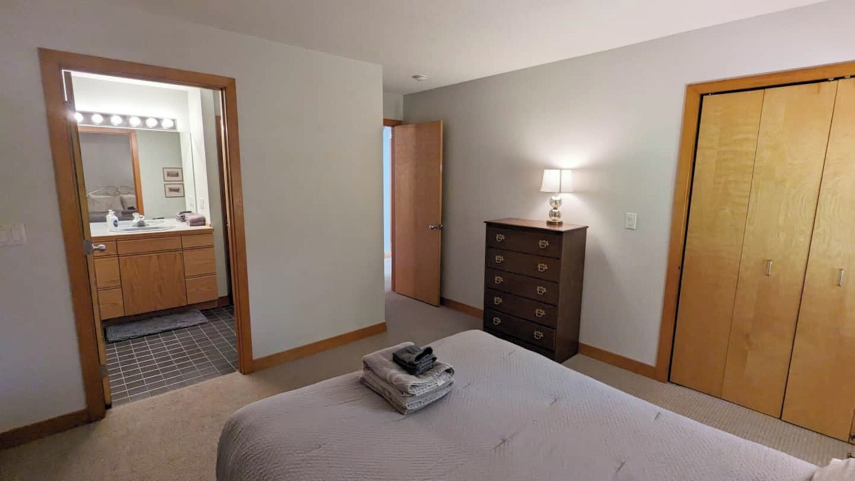A hotel room with a bed, a dresser, a mirror with lights, and a door.