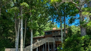 A cabin with a wooden deck and stairs leading to a balcony, surrounded by trees and a body of water. -  Temple's Sunset Harbor (Newbury)