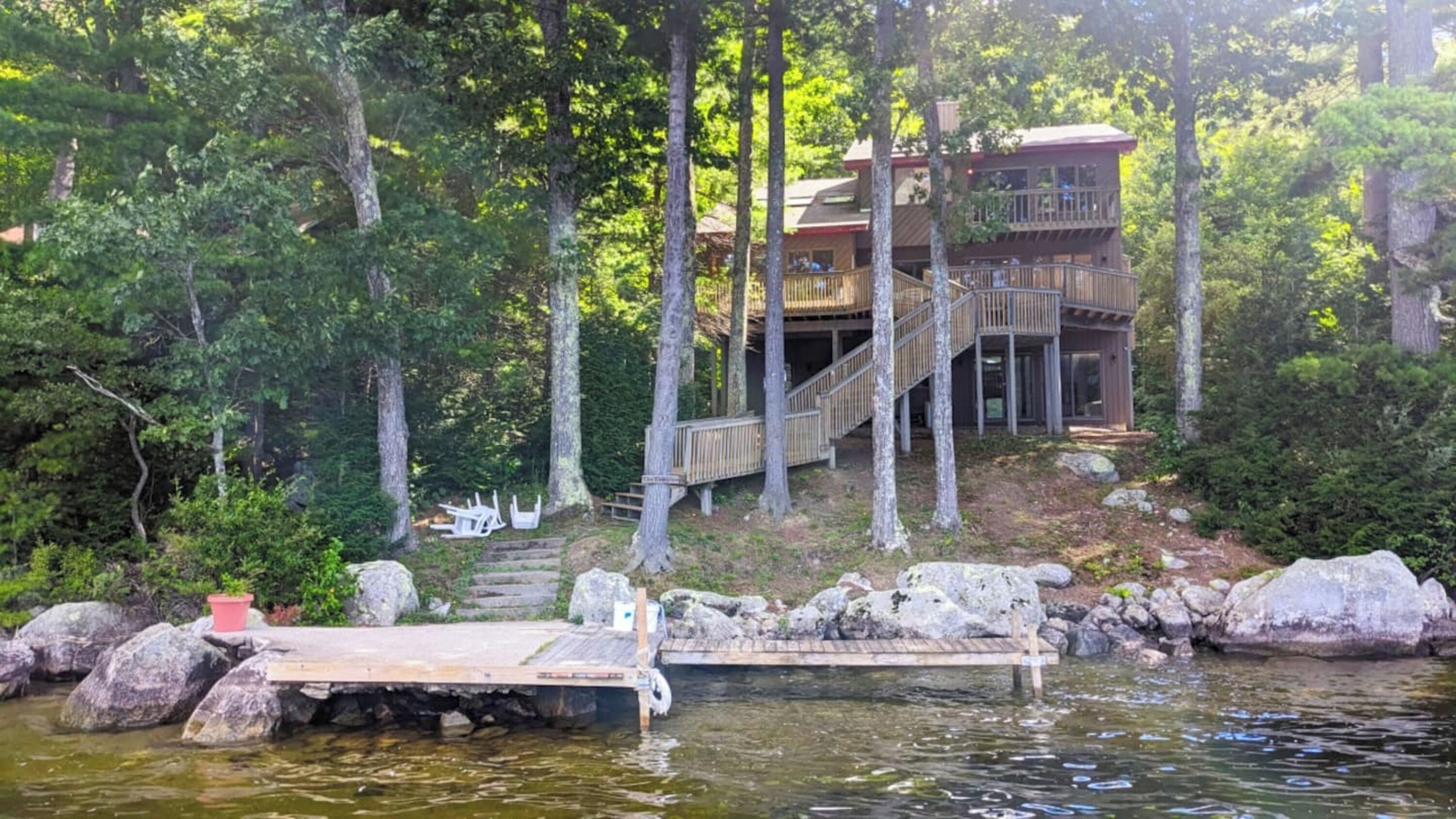 Una cabaña con terraza de madera y escaleras que conducen a un segundo nivel, rodeada de un bosque frondoso y un muelle que se adentra en un lago.
