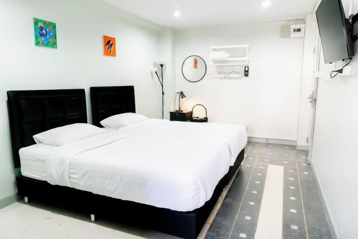 A bedroom with a double bed, a bedside table with a lamp, a wall-mounted TV, and a small window with an air conditioning unit.