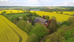 Ein Haus, umgeben von Feldern voller gelber Blumen, mit einem See und sanften Hügeln im Hintergrund.